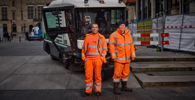 Twee mannelijke werknemers van de gemeente staan in oranje werkkleding voor een veegwagen in het centrum van Rotterdam.