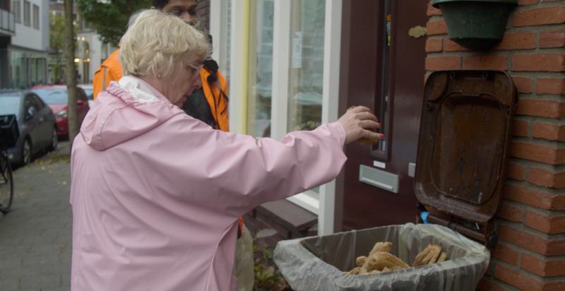 Wilma van der Wilt in een straat bij een bruine container, gooit brood in de container. Achter Wilma een gemeentemedewerker in oranje jas, kijkt toe.