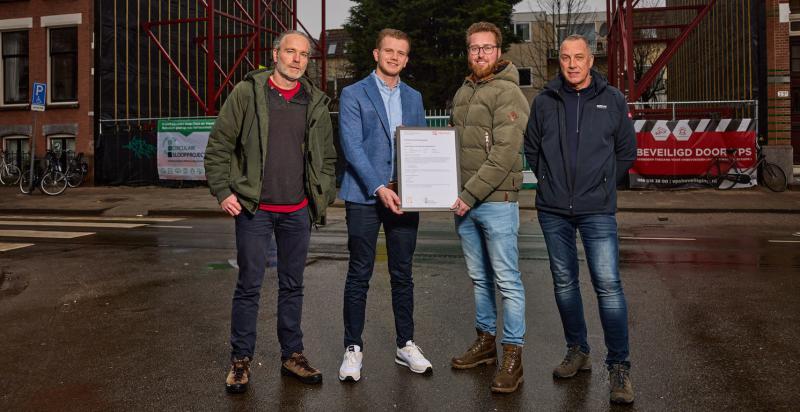 Vier mannen met op de achtergrond de bouwplaats aan de Claes de Vrieselaan. De twee mannen in het midden houden het certificaat vast.