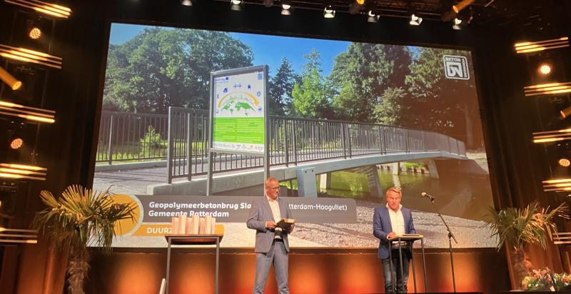 Twee mannen staan op het podium om de Betonprijs uit te reiken. Achter hen een foto van de geopolymeerbetonbrug Slaghaam.