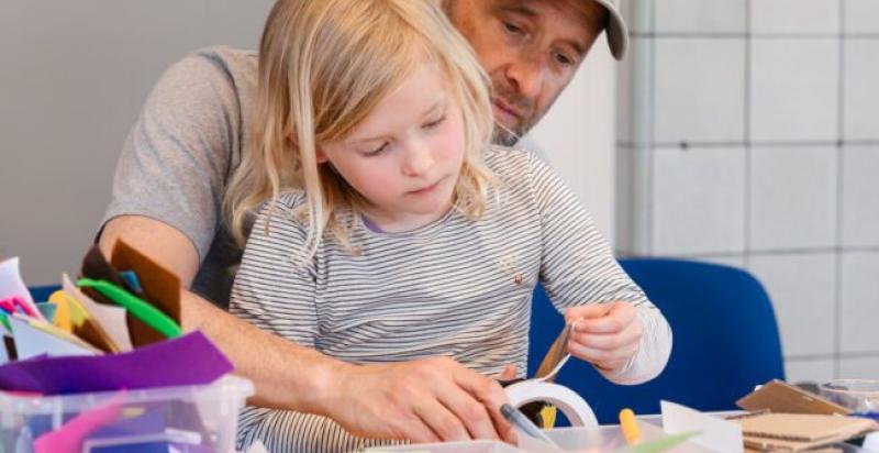 Op de foto een man met een kind, zittend aan tafel. Ze zijn samen aan het knutselen. De tafel ligt vol met kleurrijke knutselmaterialen.