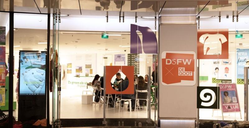ezoekers zitten aan tafels in een lichte winkelruimte tijdens een evenement van DSFW, met kleurrijke campagne-uitingen bij de ingang.