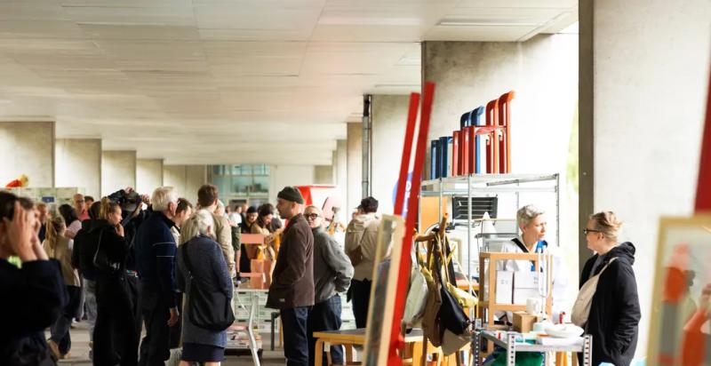 Drukke designbeurs met bezoekers die langs stands met meubels, accessoires en andere designobjecten lopen.