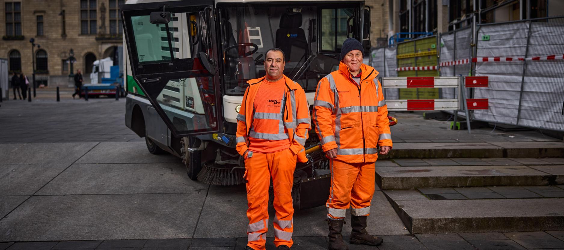 Twee mannelijke werknemers van de gemeente staan in oranje werkkleding voor een veegwagen in het centrum van Rotterdam.