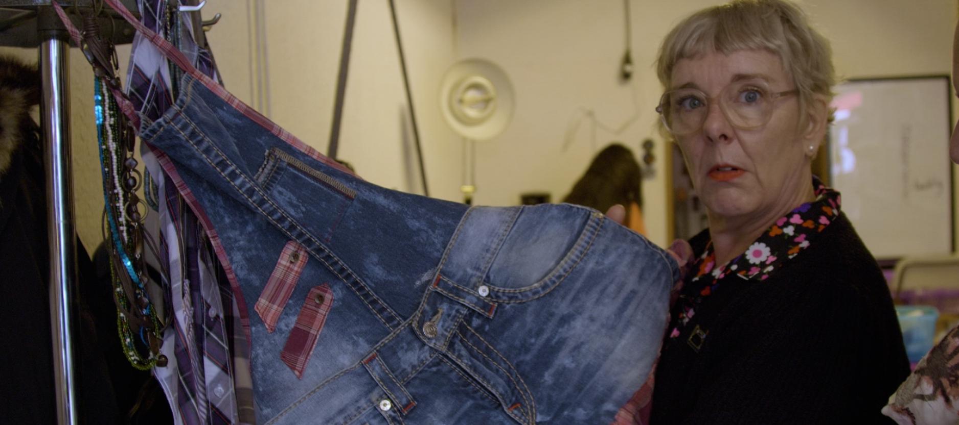 Wilma der Wilt poseert met een kledingstuk (schort van spijkerstof) in het repair atelier.
