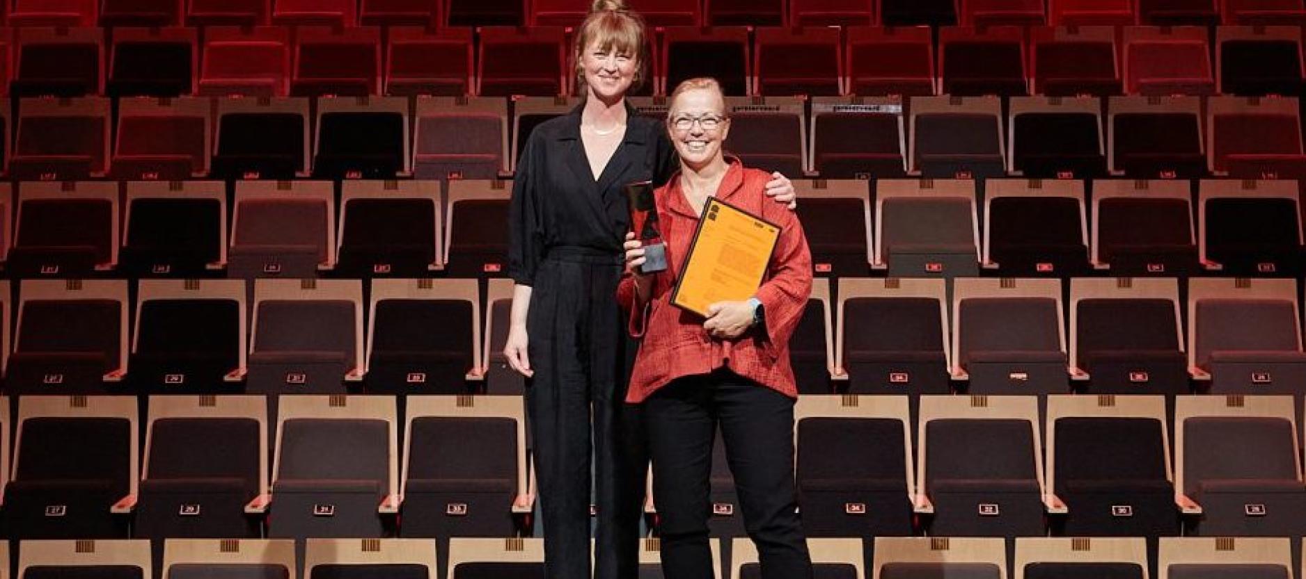 2 mensen, Christien Meindertsma en de jury, met prijs in de handen, staan voor een lege zaal op het podium.