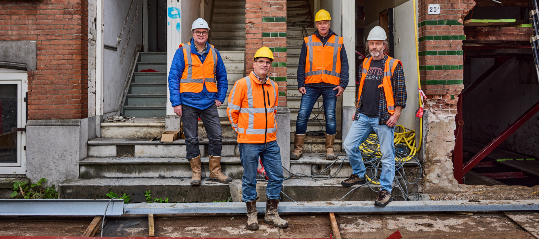 Vier mannen poseren voor een slooppand. In oranje hesjes, helm en werkschoenen.