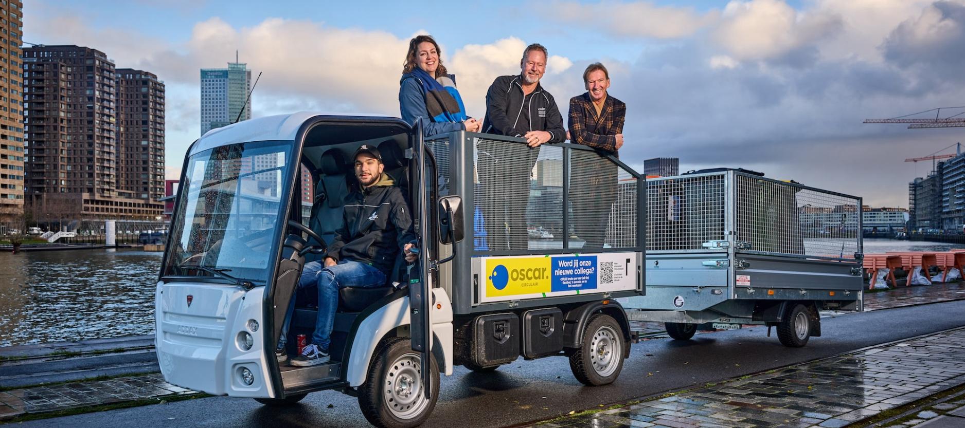 4 mensen poseren op een klein elektrisch autootje, 1 achter het stuur, 3 in de bak erachter. Auto staat langs de kade met gebouwen op de achtergrond.