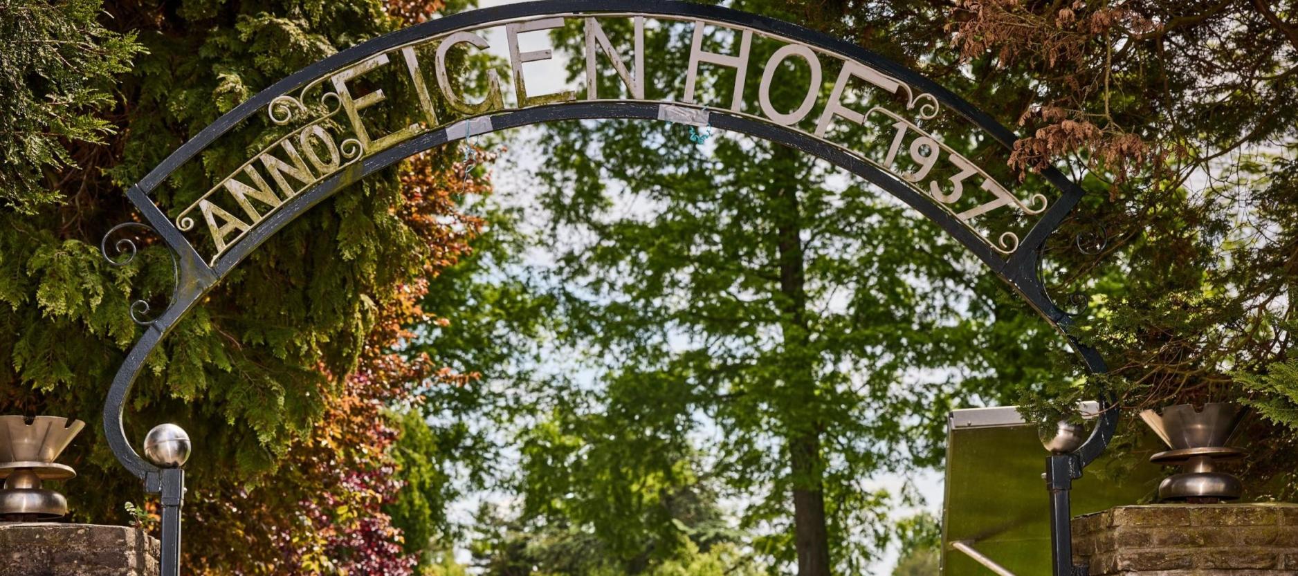 foto van mooie boog entree met de tekst "anno EIGEN HOEK 1937". De Boog is van staal op de achtergrond het groen van de stadstuin.