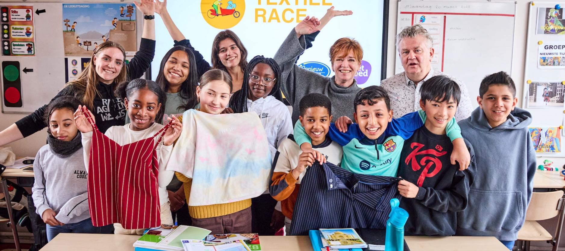 geposeerde foto van 13 vrolijke mensen, kinderen en volwassenen. De groep staat in een klaslokaal, voor het digibord met de text 'textiel race'. Kinderen houden hem, t-shirt en overhemd in hun handen.