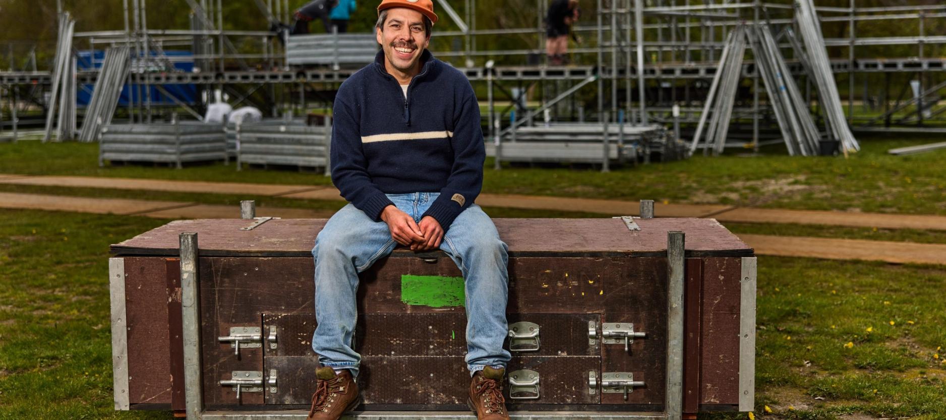 geposeerde foto van Dennis Ebeli, zit op een grote houten kist. Op de achtergrond zijn opbouw werkzaamheden te zien van een grote stellage met buizen.