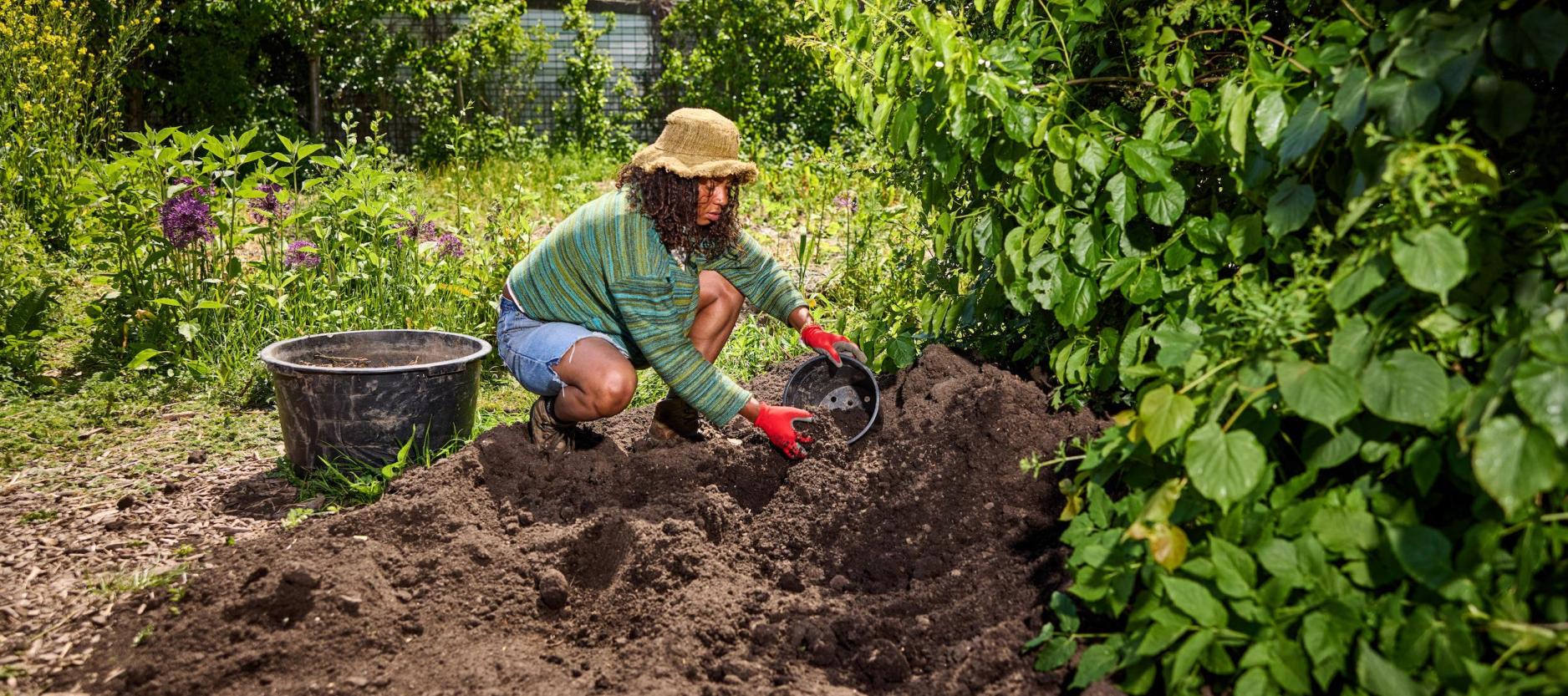 Vrouw met hoed van stro en rode handschoenen zit gehurkt op een berg aarde, tussen groene struiken en bomen. Ze is bezig aarde in een pot te stoppen.