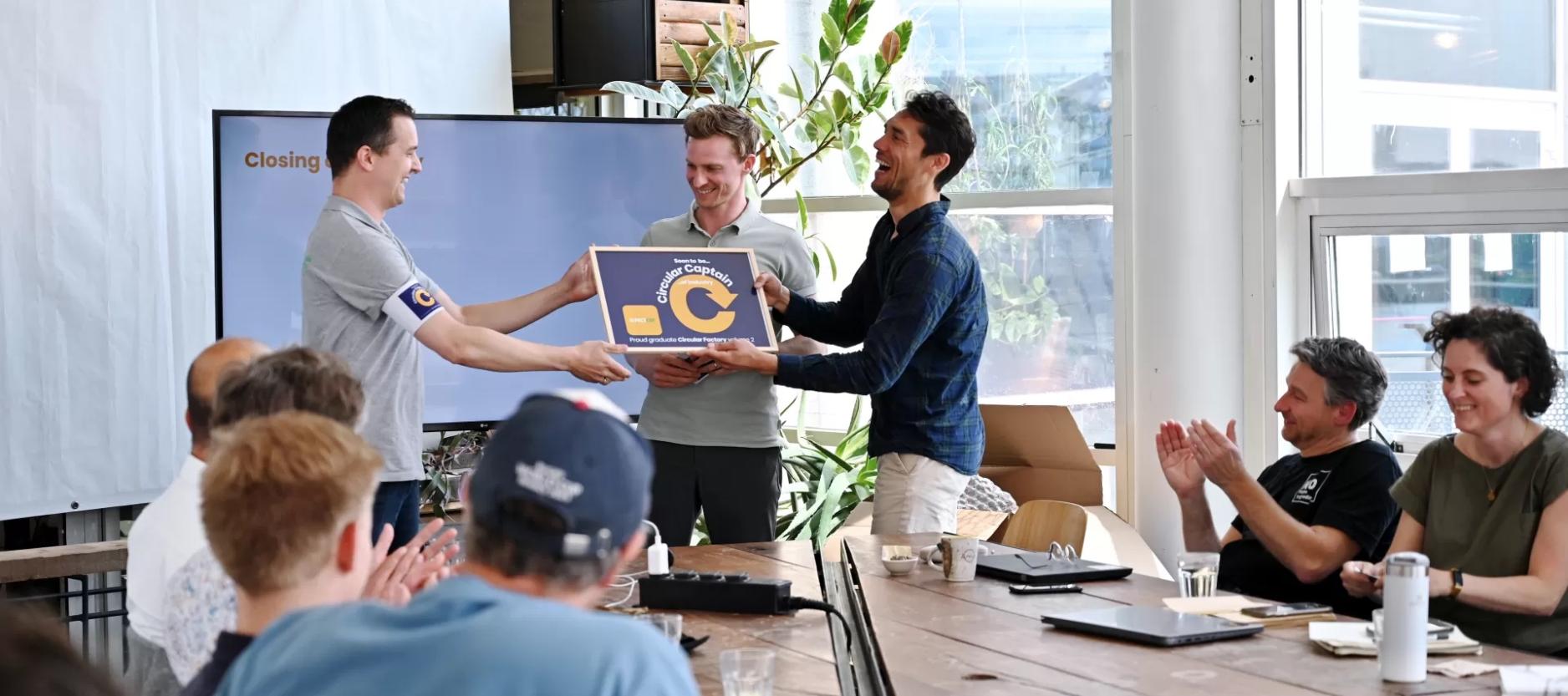 Op de foto drie mannen voor een tafel. De man links overhandigd een certificaat aan de man rechts.