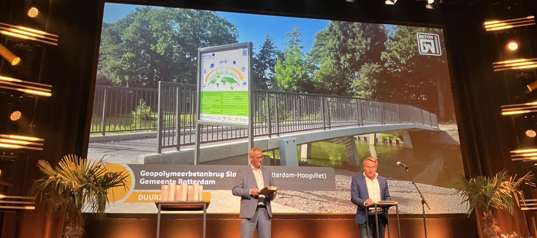 Twee mannen staan op het podium om de Betonprijs uit te reiken. Achter hen een foto van de geopolymeerbetonbrug Slaghaam.