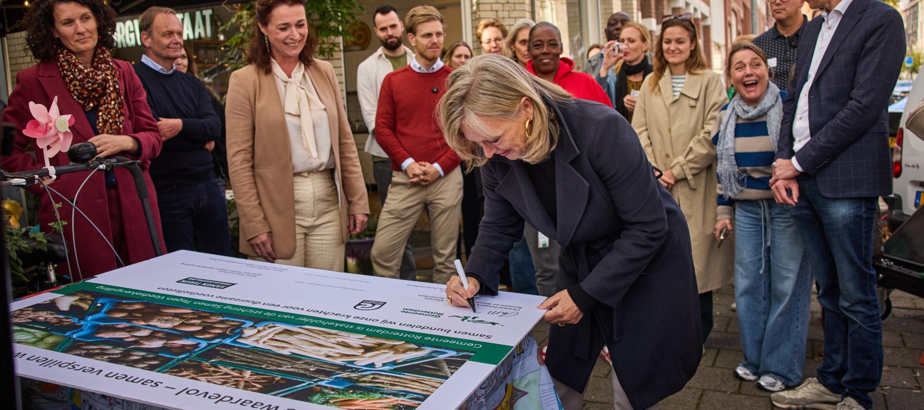 Wethouder Zeegers zet haar handtekening onder het partnerschap met stichting Samen Tegen Voedselverspilling. Een groep mensen, waaronder gedeputeerde Van Leeuwen van de Provincie Zuid-Holland kijken toe.
