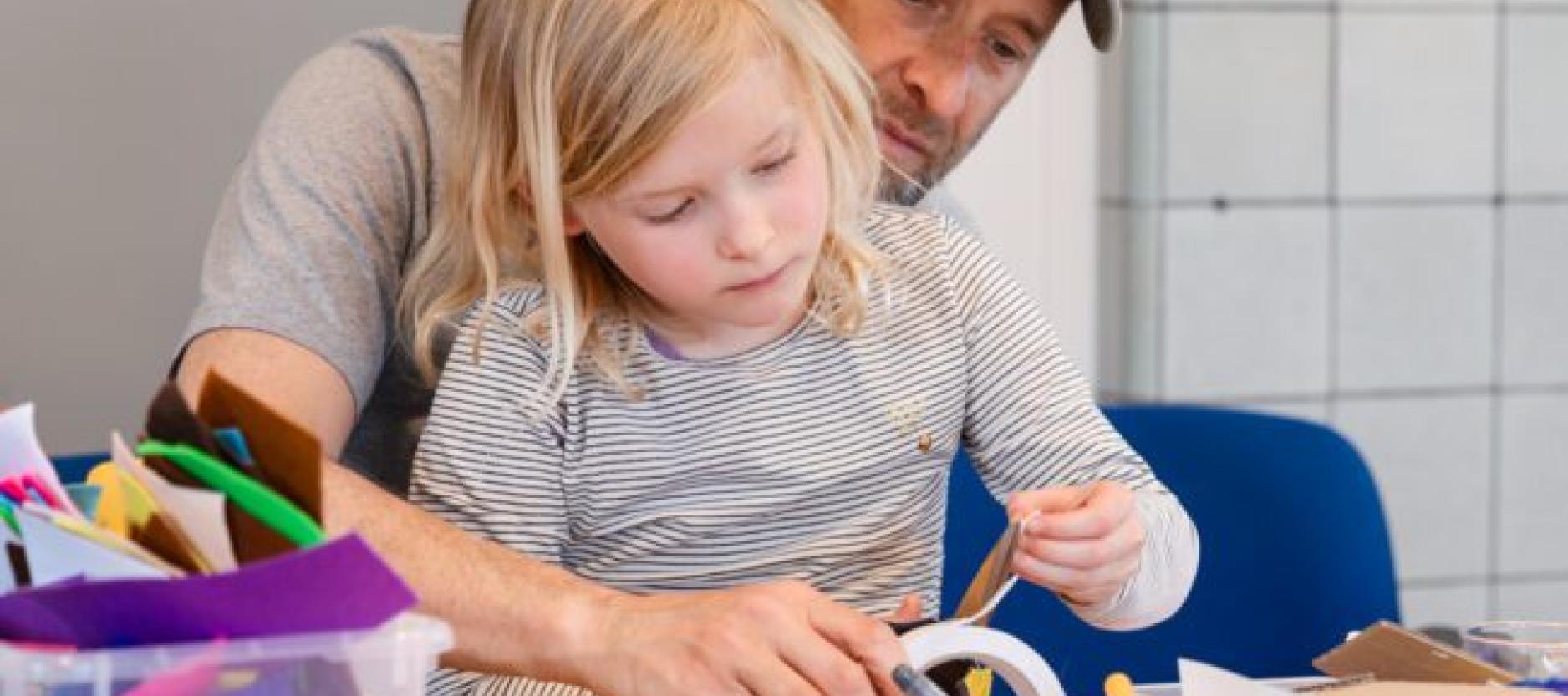 Op de foto een man met een kind, zittend aan tafel. Ze zijn samen aan het knutselen. De tafel ligt vol met kleurrijke knutselmaterialen.