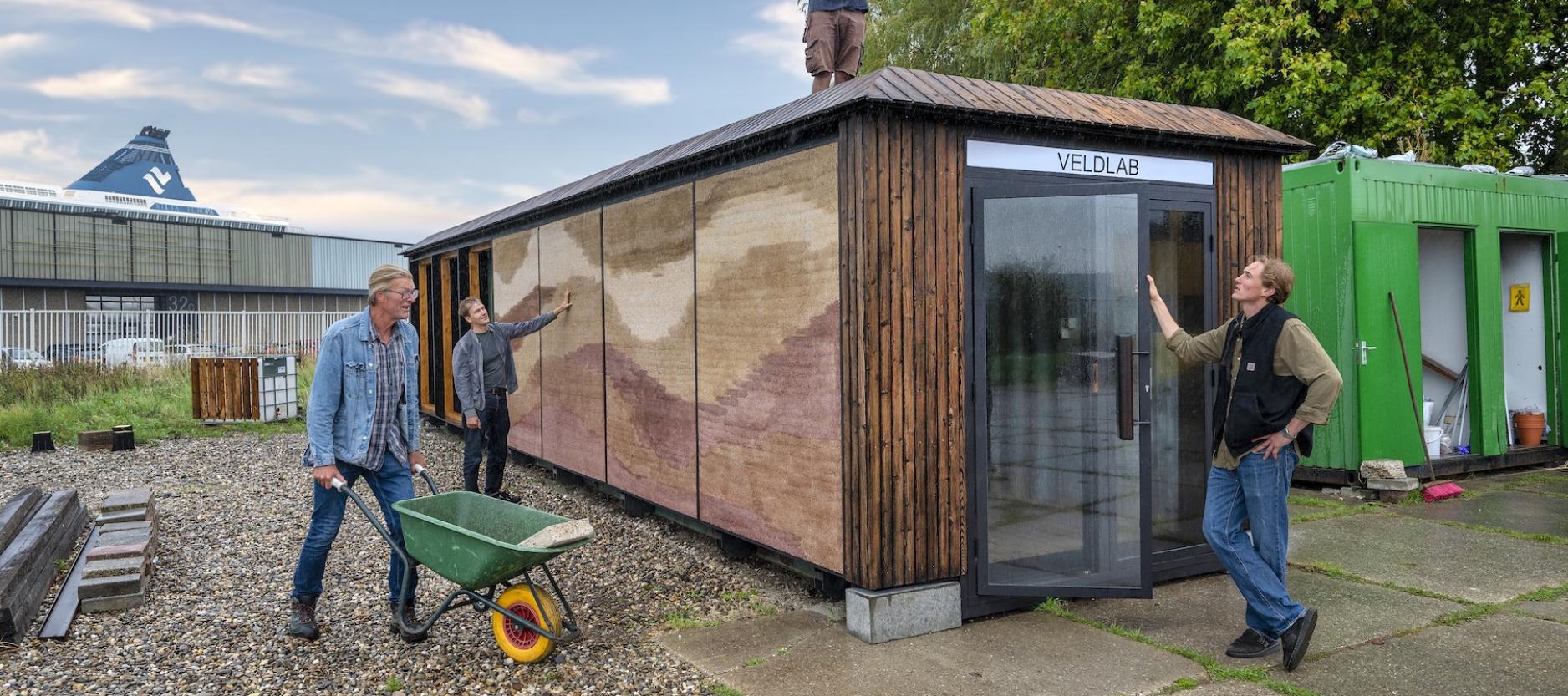 Drie mensen werken rond het Veldlab; een persoon staat op het dak en twee anderen bekijken de buitenzijde van de met natuurlijke materialen geïsoleerde zeecontainer.