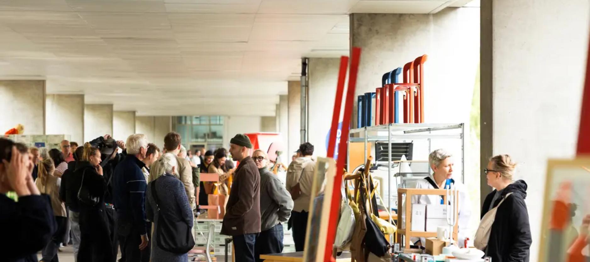 Drukke designbeurs met bezoekers die langs stands met meubels, accessoires en andere designobjecten lopen.