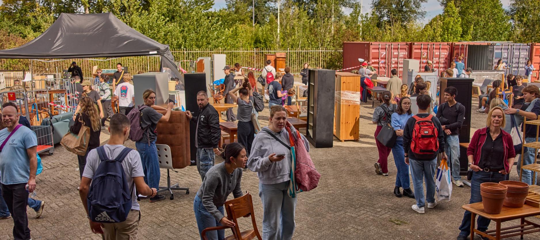 Druk buitenterrein waar bezoekers tussen uitgestalde tweedehands meubels en woonspullen rondkijken.