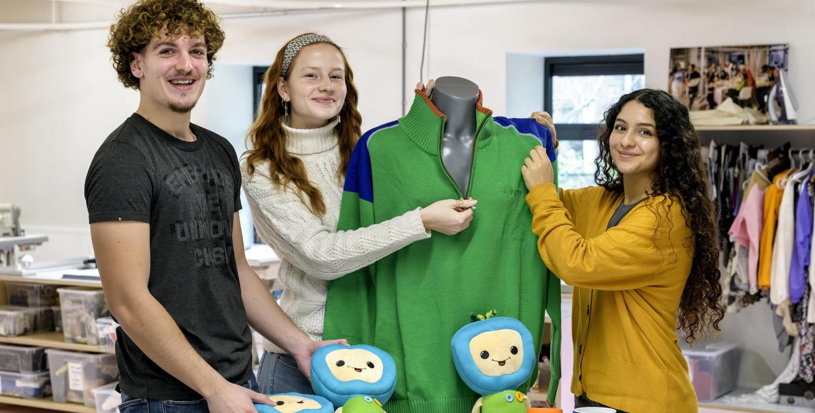 drie studenten tonen trots een groene trui en stoffen poppen.