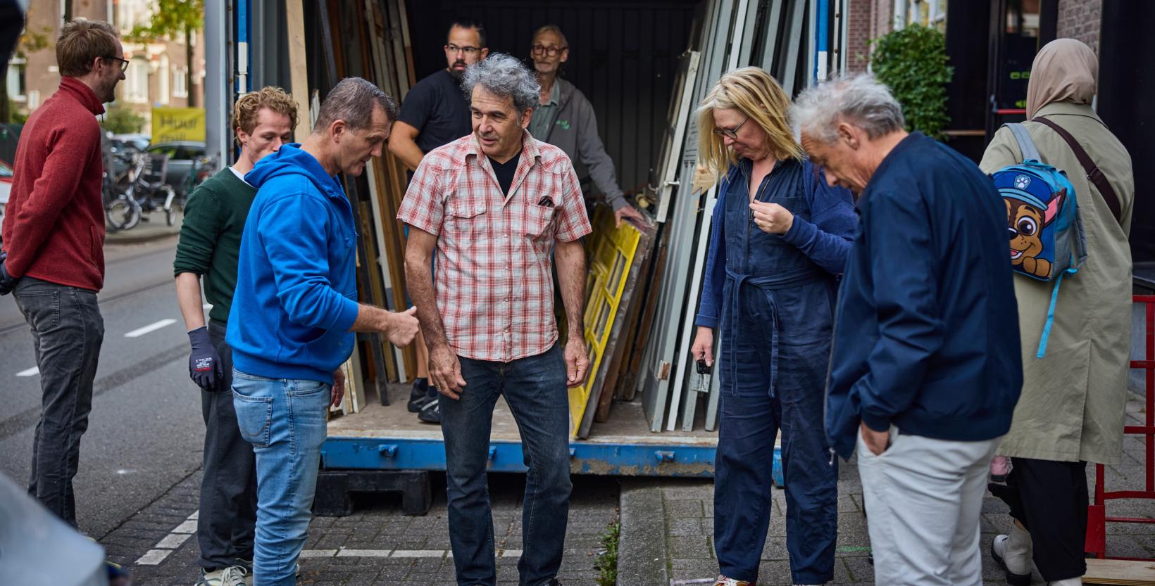 Op de foto zeven personen in de Claes de Vrieselaan. Ze staan voor een container waarin onderdelen staan die uit de sloopwoningen zijn gehaald.