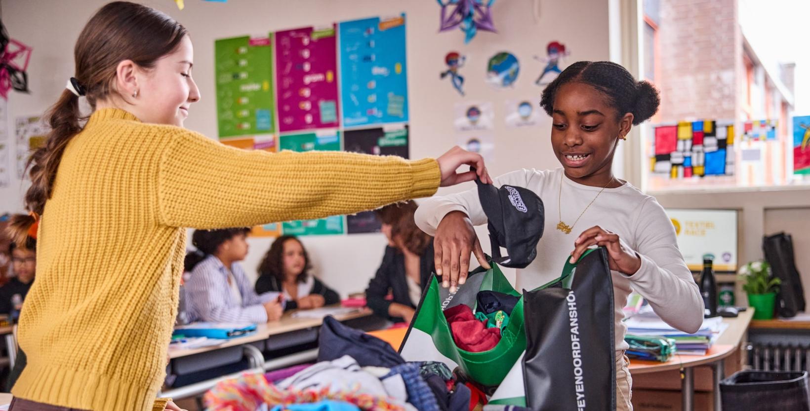 twee scholieren basisschool vullen in de klas een tas met kleding, op de foto stoppen ze net een pet in de tas.