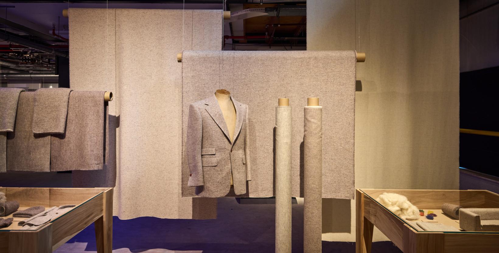 opstelling van de tentoonstelling, met grijze wollen lappen op de achtergrond, ervoor een grijs colbertjas en twee rollen met stof van grijze wol.