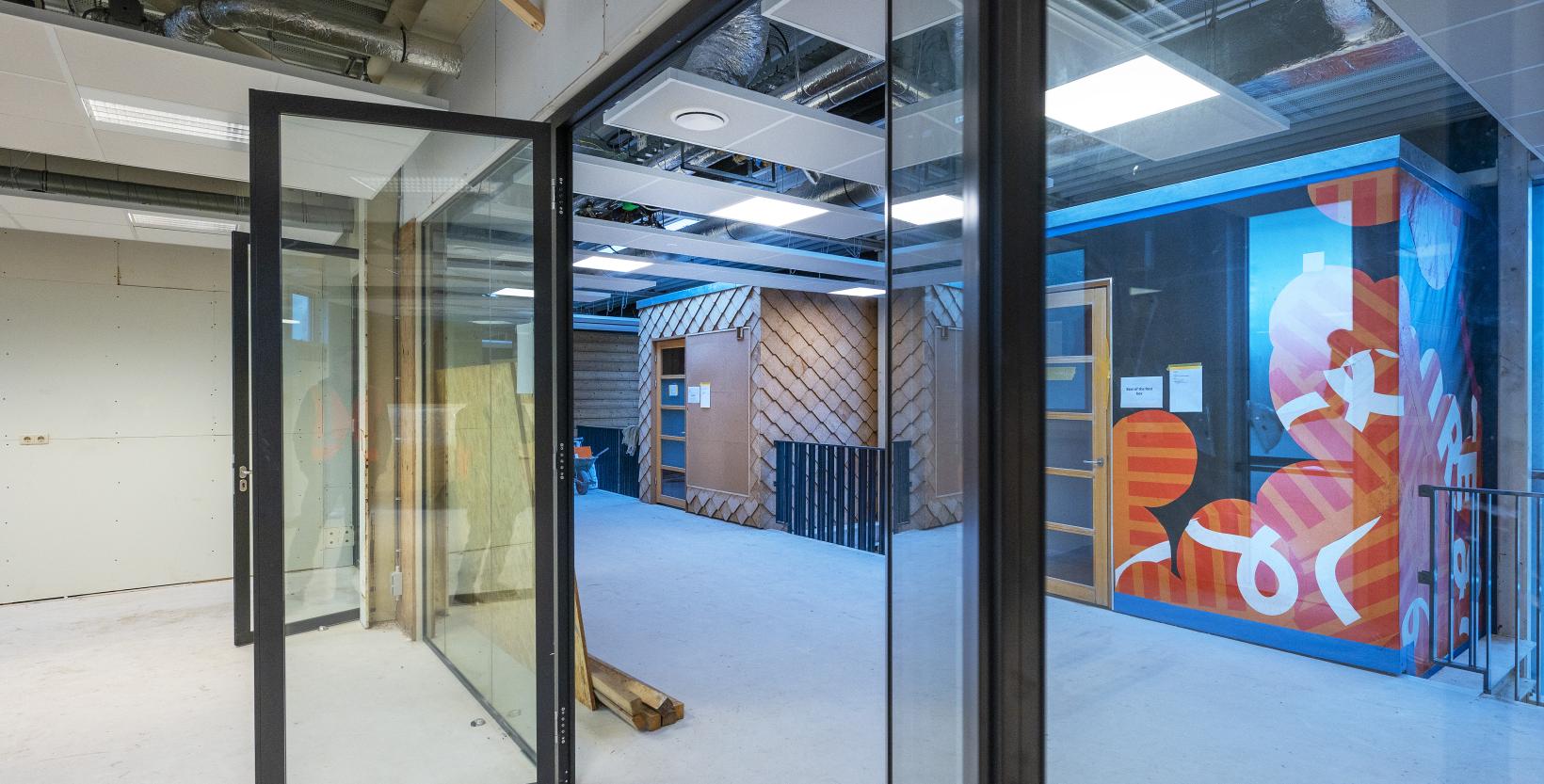 Een stukje van de binnenkant van het gebouw De HER. De foto is genomen vanuit een werkruimte met glazen wand en deur, uitkijkend op de gang.