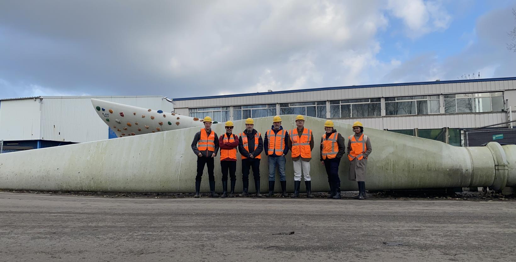 Zeven mannen staan in een oranje hesje en met bouwhelm op staan voor een grote windmolenwiek die op de grond ligt.