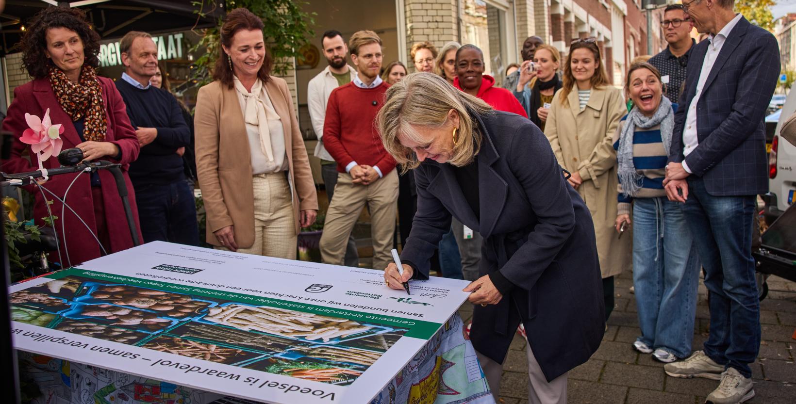 Wethouder Zeegers zet haar handtekening onder het partnerschap met stichting Samen Tegen Voedselverspilling. Een groep mensen, waaronder gedeputeerde Van Leeuwen van de Provincie Zuid-Holland kijken toe.