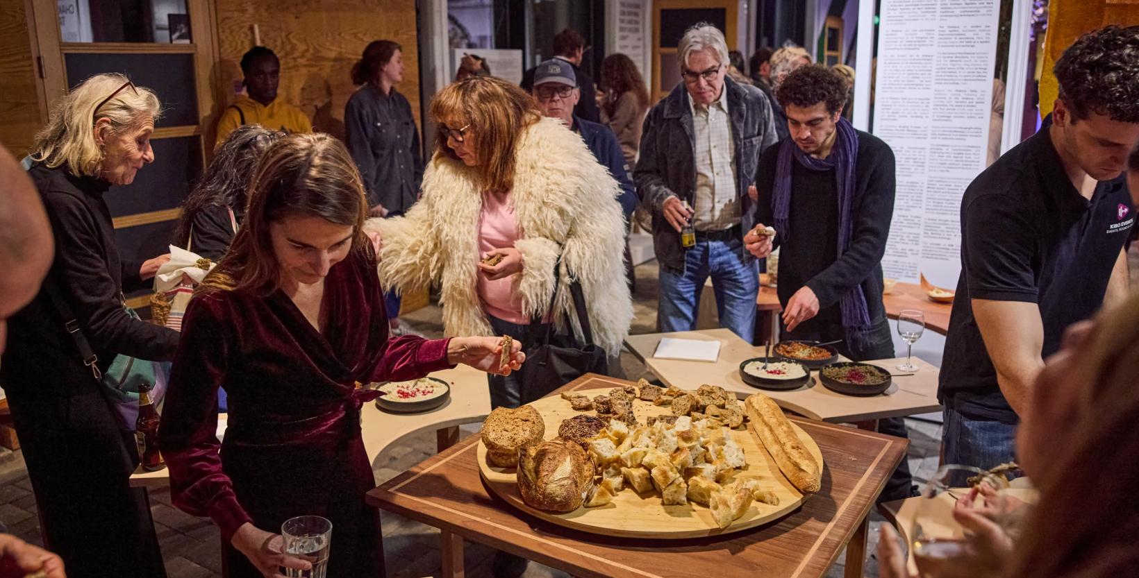 Bezoekers staan tijdens de opening van tentoonstelling AYNI in De HER rond een tafel met brood en dips.
