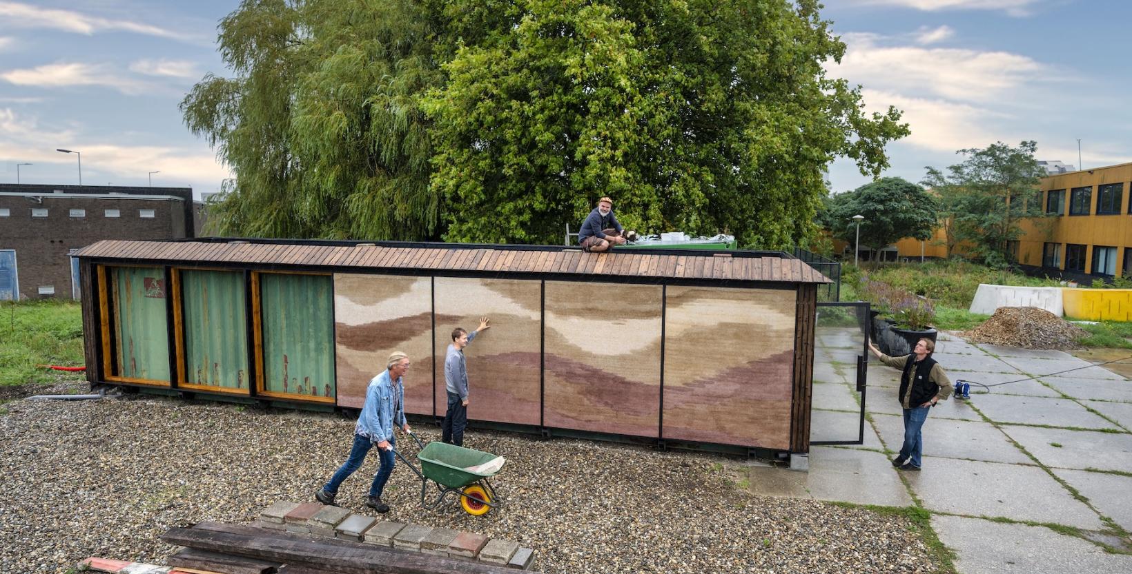Een persoon rijdt met een kruiwagen langs het Veldlab terwijl drie anderen aan de buitenkant van de zeecontainer staan.