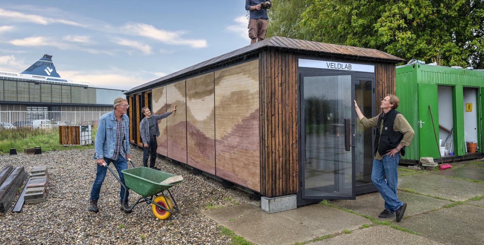 Drie mensen werken rond het Veldlab; een persoon staat op het dak en twee anderen bekijken de buitenzijde van de met natuurlijke materialen geïsoleerde zeecontainer.