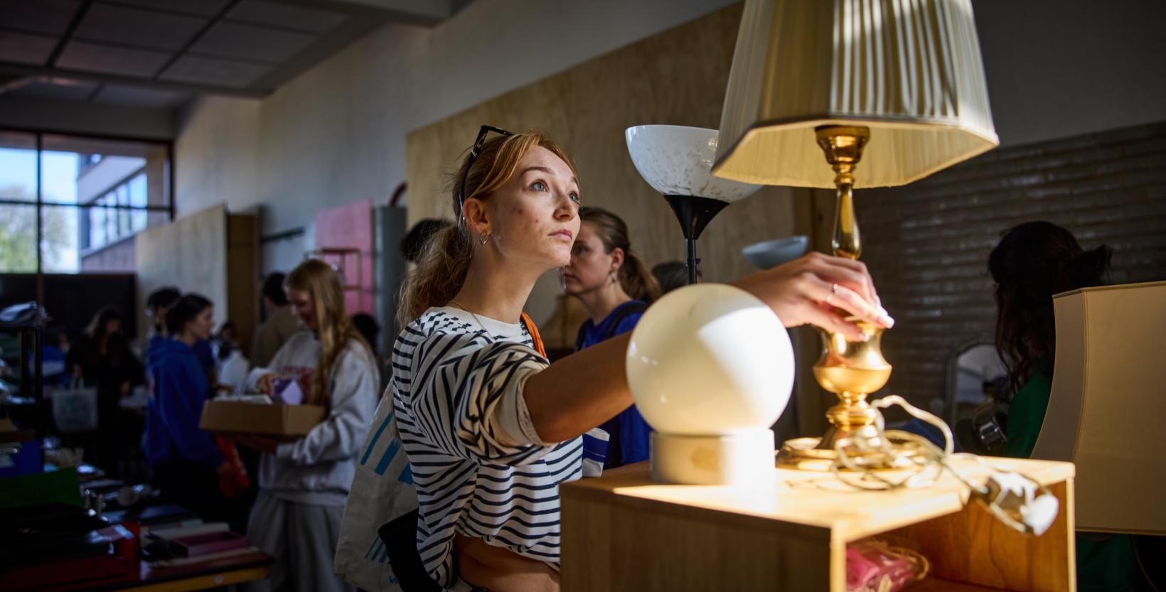 Bezoeker bekijkt een goudkleurige tafellamp tussen andere tweedehands lampen en spullen.