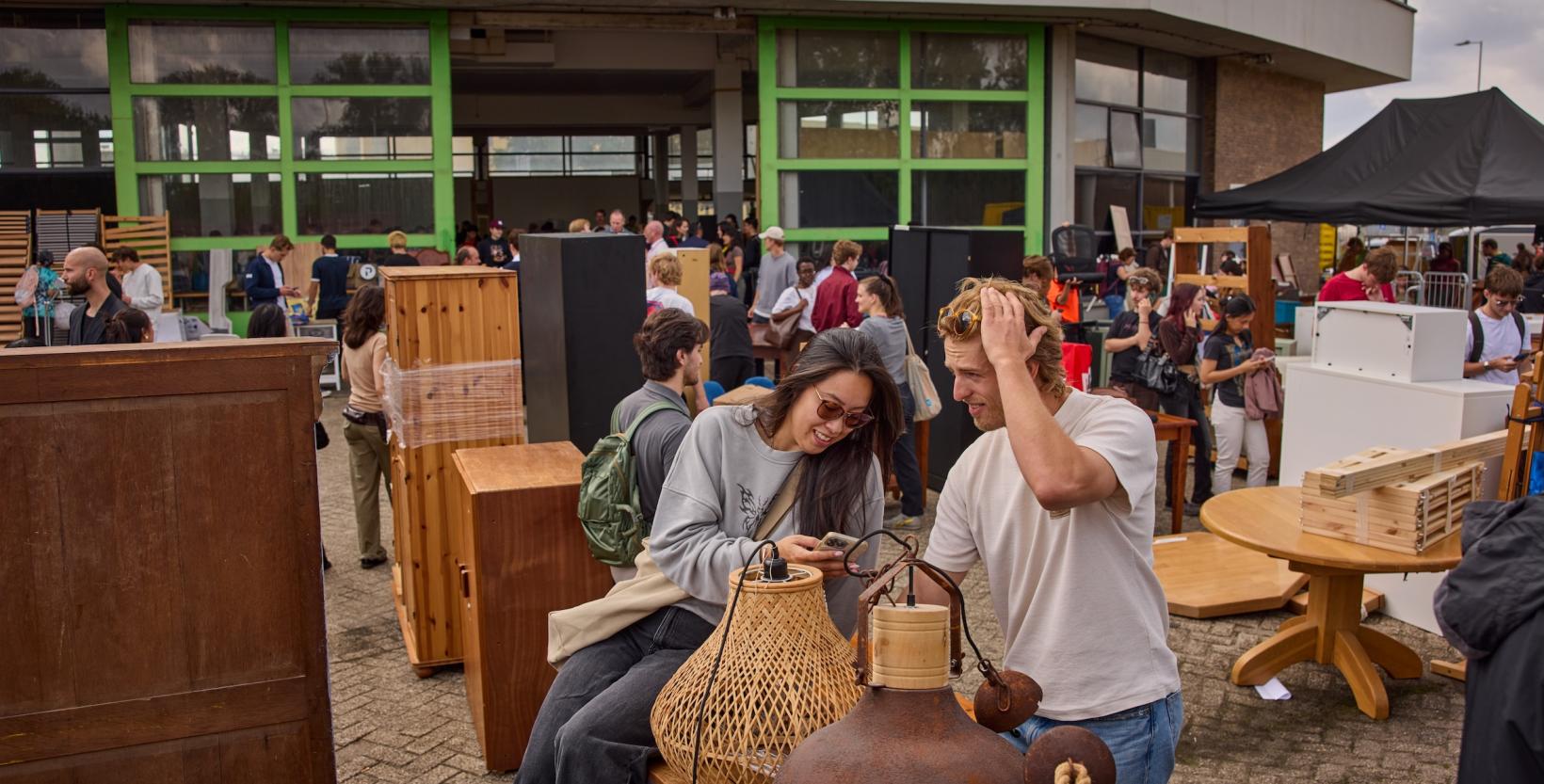 Twee bezoekers bekijken samen tweedehands lampen op een druk terrein vol meubels.