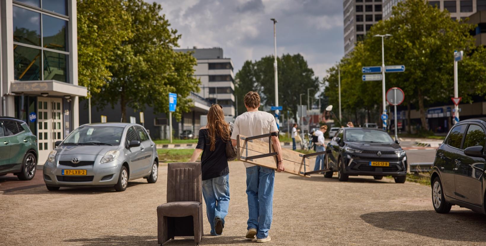 Twee bezoekers lopen met een stoel en een houten bank tussen geparkeerde auto’s.